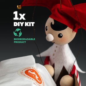 Puede incluir: Marioneta de madera hecha a mano con sombrero y atuendo de bufón rojos y blancos. La marioneta tiene una cara pintada y forma parte de un kit de bricolaje. El embalaje incluye una pegatina que dice "Hand Made Originals Czech Marionettes". También es visible el texto "Biodegradable Product".