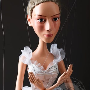 Op de afbeelding: Handgemaakte marionet van een ballerina met een serene uitdrukking. De pop draagt een witte tutu en lijfje versierd met zilveren accenten. Haar blonde haar is opgestoken in een knot en ze heeft delicate trekken. De marionet hangt aan touwtjes tegen een donkere achtergrond.
