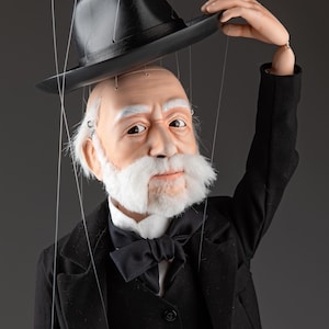 May include: A marionette puppet of an elderly person with a white mustache and hair, wearing a black suit, white collared shirt, and black bow tie. The puppet is holding a black hat above its head, with thin control strings attached to its limbs.