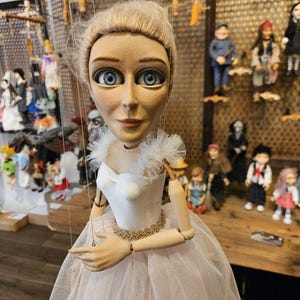 May include: A marionette puppet with blonde hair and blue eyes, wearing a white dress with a tulle skirt, ruffled shoulders, and a gold-trimmed waist. The puppet has articulated wooden arms and is suspended by thin control strings.