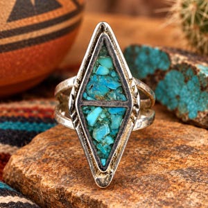 May include: A silver ring featuring a long, diamond-shaped centerpiece inlaid with crushed turquoise stone fragments. The metal frame has a dark, oxidized finish with etched detailing along the edges and a split band design on the sides.