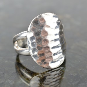May include: A silver ring with a hammered oval face. The ring has a split band design and a textured surface on the oval. The ring is reflective and appears to be made of metal.