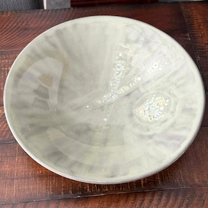 May include: A large, round, ceramic serving bowl with a pale green glaze. The bowl has a wide rim and a shallow depth. The interior features subtle, swirling patterns and decorative accents.