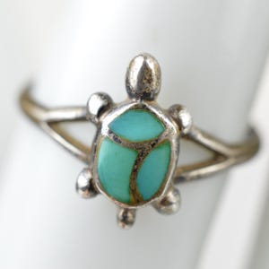 May include: A silver-toned ring featuring a turtle-shaped centerpiece. The shell is inlaid with three smooth, light blue-green turquoise-colored stones separated by thin silver lines. The band splits into two thin arms that connect to the turtle body.