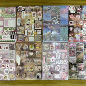May include: A collection of sticker sheets featuring various designs. The sheets include stickers with images of bears, Hello Kitty, and landscape scenes. The stickers are in various colors, including pink, white, and blue.