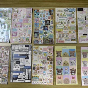 May include: A collection of sticker sheets featuring various cartoon characters and designs. The stickers include images of animals, food, and text bubbles in a variety of colors. The sheets are arranged on a wooden surface.