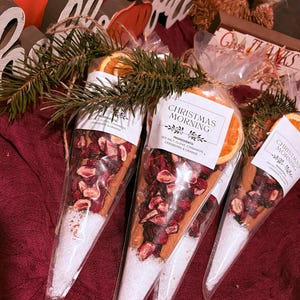 May include: Three cone-shaped clear plastic bags filled with layers of sea salt, dried cranberries, cinnamon sticks, and dried orange slices. Each bag features a white label with the text CHRISTMAS MORNING and a list of ingredients, tied with twine and a small pine sprig.