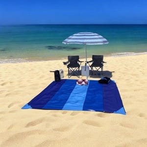 May include: A beach scene with a blue and white striped umbrella, two black folding chairs, and a blue beach blanket. A cooler, a small speaker, and a folded towel are also on the blanket. The beach is sandy, and the ocean is in the background.