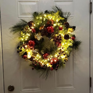 May include: A festive Christmas wreath with a circular design, adorned with red ornaments, pinecones, and berries. The wreath is illuminated by warm white fairy lights, creating a glowing effect. The wreath is displayed on a white door.