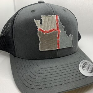 May include: A grey trucker cap with a black mesh back and a curved peak. The cap features an embroidered patch in the shape of a state, with red and white stitching details. It has a snapback closure.