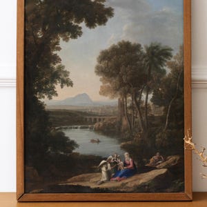 May include: A framed landscape painting with a wooden frame. The artwork depicts a river scene with trees, mountains, and figures. The painting is displayed on a wooden cabinet, with a decorative branch in the foreground.