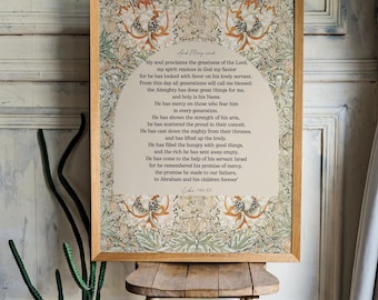 May include: A framed print featuring a floral design with text from Luke 1:48-55. The print is displayed on a small wooden stool. The frame is light brown, and the print has a beige background with a floral pattern in shades of orange, green, and brown.