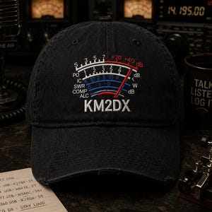 May include: Black distressed baseball cap with an embroidered graphic of a radio frequency meter. The meter displays numbers and the text "KM2DX". A notepad, pen, and a coaster with the text "GOOD SIGNALS GOOD PEOPLE" are also visible.
