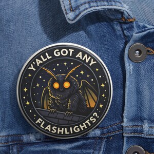 Pode incluir: Um botão redondo preto com uma ilustração de desenho animado de uma mariposa. A mariposa tem olhos laranja e está pousada num telhado. O texto no botão diz "Y'all Got Any Flashlights?" em letras brancas, rodeado por estrelas.