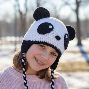 May include: A white and black crochet panda hat with ear flaps and braided ties. The hat features black panda ears and eye patches. The hat is worn by a person wearing a pink sweater.