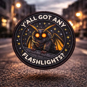 Op de afbeelding: Ronde zwarte patch met een cartoon mot ontwerp, met gloeiende oranje ogen en vleugels. De patch heeft de tekst "Y'ALL GOT ANY FLASHLIGHTS?" in het wit, omringd door sterren.