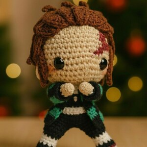 May include: A crocheted doll depicting a character with brown hair and a forehead scar. The doll is dressed in a black and green outfit with white details. It has large black eyes and is posed with hands clasped together.