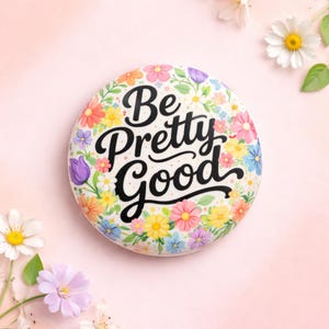 Pode incluir: Um botão redondo com um padrão floral e a frase "Be Pretty Good" em preto. As flores são rosa, amarelas, azuis e roxas sobre um fundo branco. O fundo é rosa claro, criando um ambiente alegre e primaveril.