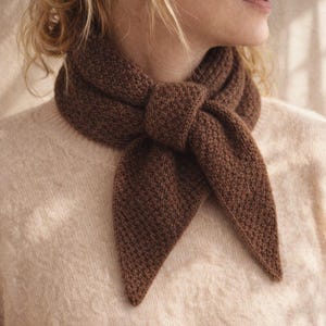 Puede incluir: Bufanda marrón tejida anudada. La bufanda está hecha de un hilo marrón oscuro texturizado y está drapeada alrededor del cuello. Los extremos de la bufanda son puntiagudos. El fondo es de color beige claro.