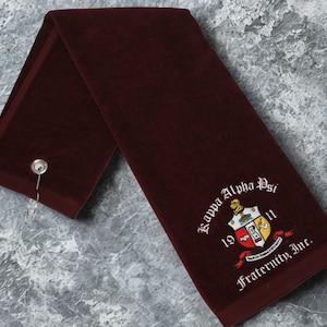 May include: A maroon towel with the embroidered text "Kappa Alpha Psi Fraternity, Inc." and a crest. The towel has a metal grommet and a silver hook. The towel is folded and lies on a gray surface.