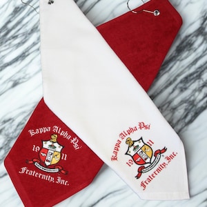 May include: Two golf towels, one red and one white, with a shield emblem and the text "Kappa Alpha Psi Fraternity, Inc." embroidered in red and black. Each towel has a metal ring for hanging.