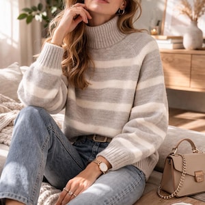 Puede incluir: Un jersey de cuello alto a rayas grises y blancas con mangas largas. El jersey se combina con vaqueros azul claro. También se ve un bolso beige con una correa de cadena dorada y un reloj dorado.