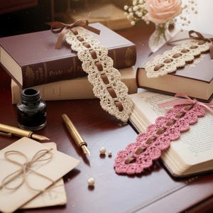 Puede incluir: Marcadores de ganchillo hechos a mano en crema y rosa, adornados con cintas marrones. Los marcadores descansan sobre libros abiertos y un escritorio de madera, junto con un tintero, plumas y papelería, creando una estética vintage.