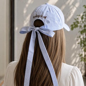May include: A light blue baseball cap with thin white vertical stripes. The back features an adjustable opening tied with a matching fabric ribbon bow. Embroidered in brown thread above the opening is the text today is ok.