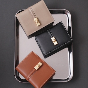 Puede incluir: Tres carteras de cuero en beige, negro y marrón, cada una con un cierre dorado y un diseño cosido. Las carteras, de forma cuadrada, descansan sobre una bandeja plateada, ofreciendo un accesorio clásico y elegante.
