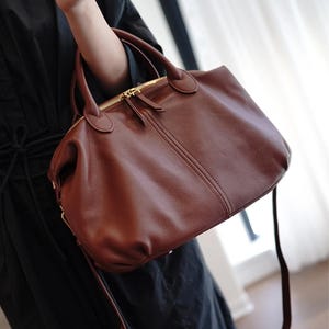 Puede incluir: Un bolso de mano de cuero marr&oacute;n con una asa superior y una correa larga para el hombro. El bolso tiene un cierre de cremallera dorado y un peque&ntilde;o tirador de cremallera. El bolso lo sostiene una persona que lleva un vestido negro.