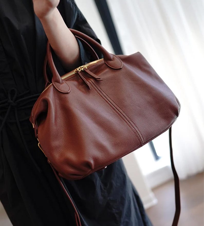 Puede incluir: Un bolso de mano de cuero marr&oacute;n con una asa superior y una correa larga para el hombro. El bolso tiene un cierre de cremallera dorado y un peque&ntilde;o tirador de cremallera. El bolso lo sostiene una persona que lleva un vestido negro.
