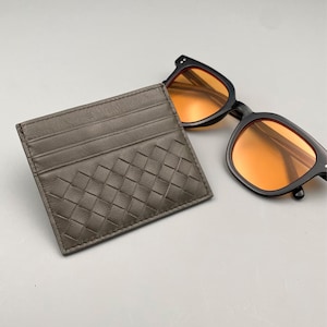 May include: A grey leather cardholder with a woven design and multiple card slots. Beside it are black sunglasses with orange lenses. The cardholder and sunglasses are on a grey surface.