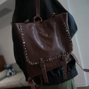 Puede incluir: Una mochila de cuero marrón oscuro con una solapa plegable asegurada con dos correas con hebilla. Los bordes de la solapa están decorados con una hilera de tachuelas metálicas intercaladas con pequeños detalles decorativos redondos en azul claro y rojo oscuro.