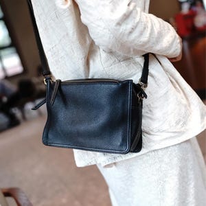 May include: A black leather crossbody bag with a rectangular shape and a zip closure. The bag has a long, adjustable strap and is worn over the shoulder. The bag is made of a textured leather material.