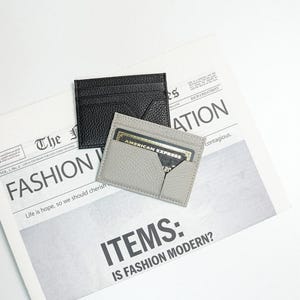 May include: Two leather cardholders, one black and one grey, sit on a newspaper with the headline "FASHION". The grey cardholder contains a credit card. The newspaper's text includes "ITEMS: IS FASHION MODERN?" and "AMERICAN EXPRESS".