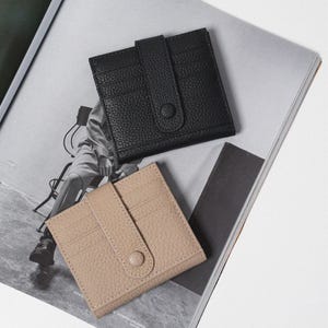 Puede incluir: Dos tarjeteros de cuero, uno negro y otro beige, se muestran sobre una revista. Cada tarjetero tiene múltiples ranuras para tarjetas y un cierre de solapa con un botón. Los tarjeteros son rectangulares y tienen una superficie texturizada.