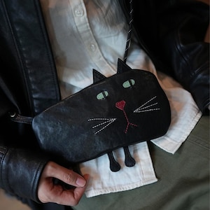 Könnte beinhalten: Schwarze Leder-Umhängetasche in Form einer Katze. Die Tasche hat grüne Augen, eine rote Nase und weiße, aufgestickte Schnurrhaare. Die Ohren und Beine der Katze sind ebenfalls schwarz. Sie hat einen schwarzen Riemen.