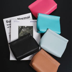 Puede incluir: Una colección de pequeños monederos de colores rosa, turquesa, azul claro, marrón y negro. Los monederos están hechos de material texturizado y tienen un diseño rectangular simple. Se ve una revista con la palabra "Normal".