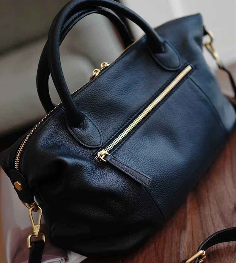 Puede incluir: Bolso de mano de cuero negro con cremalleras y herrajes dorados. El bolso tiene dos asas y una correa para el hombro desmontable. El cuero tiene un aspecto texturizado. El bolso est&aacute; sobre una superficie de madera.