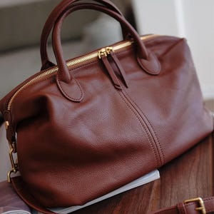 Puede incluir: Un bolso de mano de cuero marr&oacute;n con cierre de cremallera superior y dos asas. El bolso tiene una correa para el hombro desmontable y herrajes dorados. El bolso est&aacute; sobre una superficie de madera.
