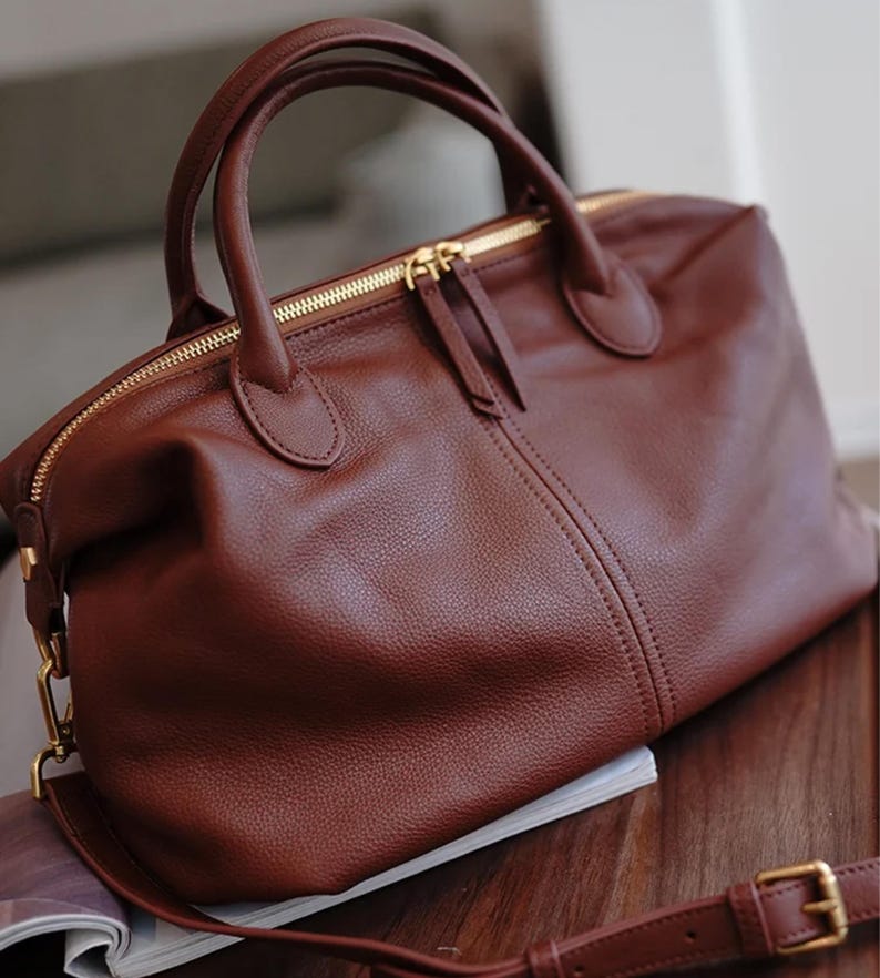 Puede incluir: Un bolso de mano de cuero marr&oacute;n con cierre de cremallera superior y dos asas. El bolso tiene una correa para el hombro desmontable y herrajes dorados. El bolso est&aacute; sobre una superficie de madera.