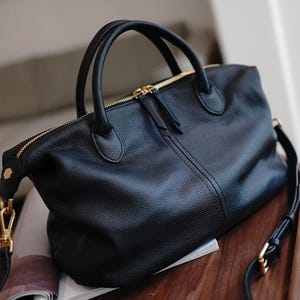 Puede incluir: Bolso de mano de cuero negro con herrajes dorados. El bolso tiene cierre de cremallera superior, dos asas superiores y una correa de hombro desmontable. El cuero tiene una textura. El bolso est&aacute; sobre una revista.