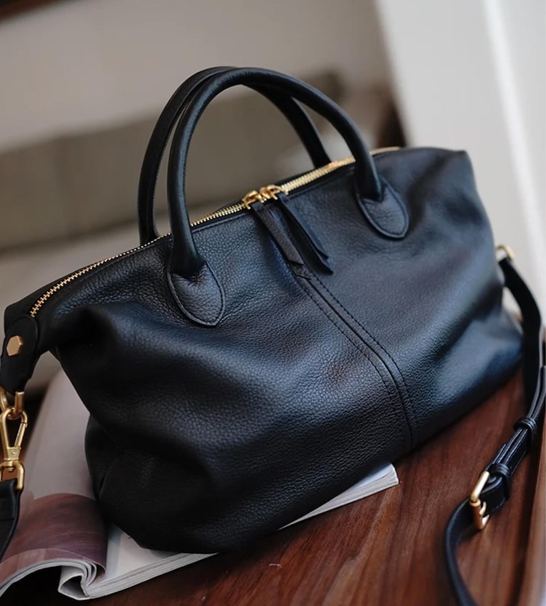 Puede incluir: Bolso de mano de cuero negro con herrajes dorados. El bolso tiene cierre de cremallera superior, dos asas superiores y una correa de hombro desmontable. El cuero tiene una textura. El bolso est&aacute; sobre una revista.