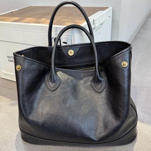 Könnte beinhalten: Eine schwarze Ledertasche mit zwei Henkeln und goldfarbener Hardware. Die Tasche hat eine rechteckige Form mit flachem Boden und einer Innentasche mit Reißverschluss. Die Tasche ist oben offen und scheint leer zu sein.