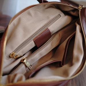 Puede incluir: Vista interior de un bolso de mano de cuero marr&oacute;n. El bolso tiene m&uacute;ltiples compartimentos con cremallera y herrajes dorados. El forro es de tela beige claro y se ve una peque&ntilde;a etiqueta de cuero marr&oacute;n.