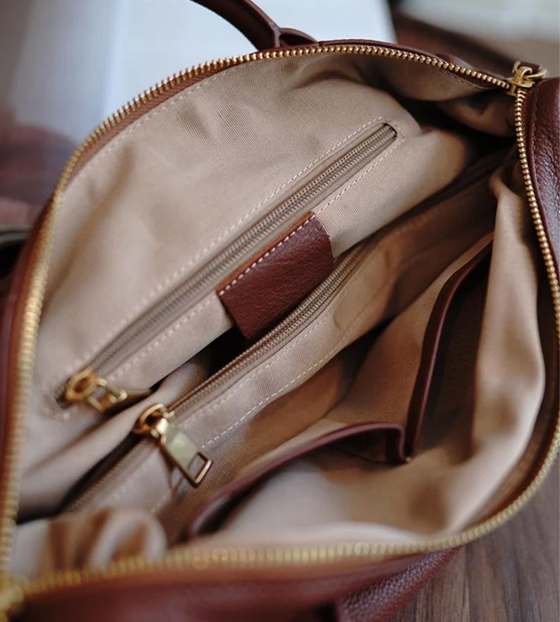 Puede incluir: Vista interior de un bolso de mano de cuero marr&oacute;n. El bolso tiene m&uacute;ltiples compartimentos con cremallera y herrajes dorados. El forro es de tela beige claro y se ve una peque&ntilde;a etiqueta de cuero marr&oacute;n.