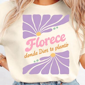 Puede incluir: Camiseta color crema con un diseño floral y el texto "Florece donde Dios te plantó" en rosa y naranja. Las flores son moradas con centros amarillos y detalles verdes. La camiseta es una prenda casual.