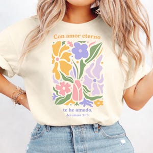 May include: Cream-colored t-shirt featuring a floral design in pastel colors. The design includes flowers in shades of yellow, purple, pink, and green, with the text "Con amor eterno te he amado. Jeremias 31:3" in a matching color scheme.