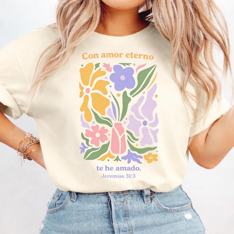 May include: Cream-colored t-shirt featuring a floral design in pastel colors. The design includes flowers in shades of yellow, purple, pink, and green, with the text "Con amor eterno te he amado. Jeremias 31:3" in a matching color scheme.