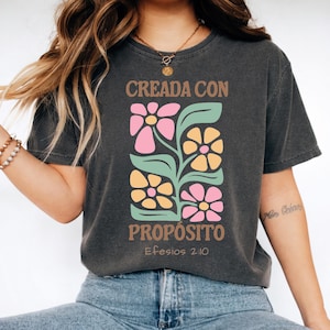 May include: Dark gray t-shirt with a floral design in pink, yellow, and green. The shirt features the text "CREADA CON PROP&Oacute;SITO" and "Efesios 2:10" in brown. The design is centered on the front of the shirt.